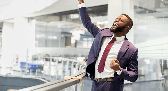 Cheerful african american young businessman celebrating success, handsome black manager in suit and tie raising hands up and screaming, office building interior, panorama with copy space