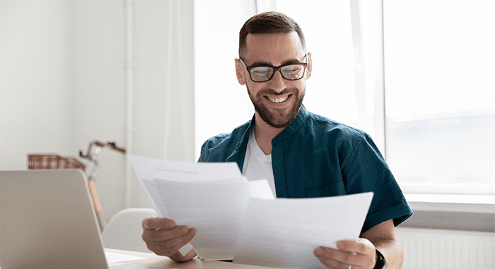 Happy young businessman in glasses looking through paper documents, satisfied with research results. Smiling male accounting manager reviewing financial report, sitting at table alone at office.