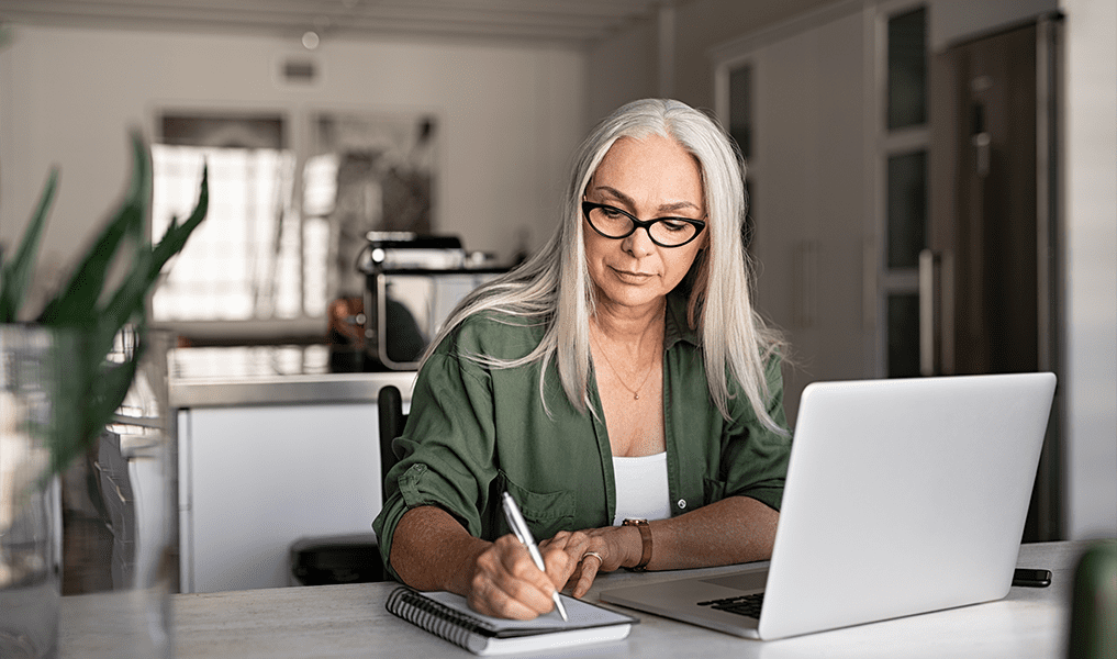 Senior stylish woman taking notes in notebook while using laptop at home. Old freelancer writing details on book while working on laptop in living room. Focused cool lady writing notary in notepad.